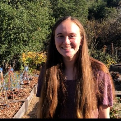 young woman, Kiera H., smiling. Behind her is a garden bed and trees.
